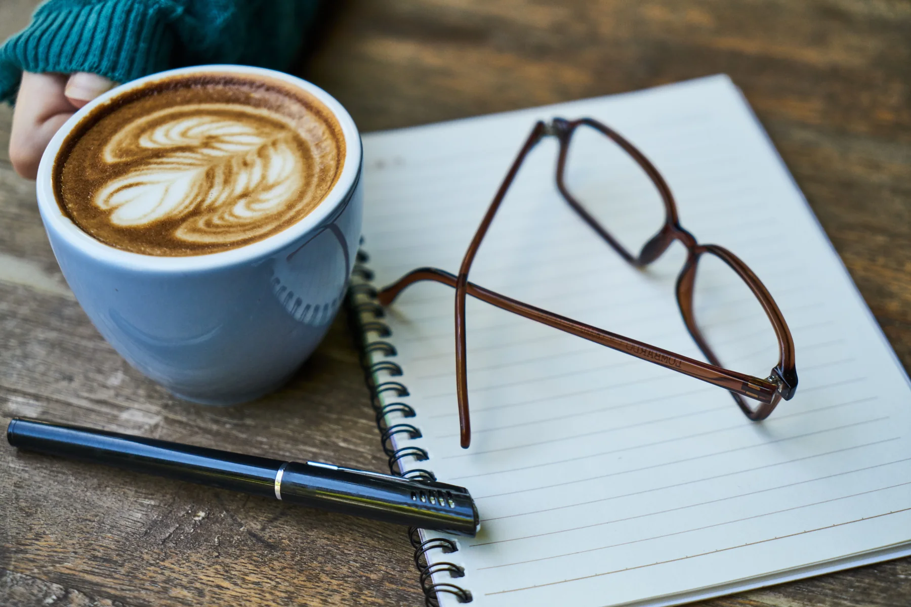 Eine Tasse Milchkaffee, eine Lesebrille und ein Füller auf einem aufgeschlagenen Notizbuch.