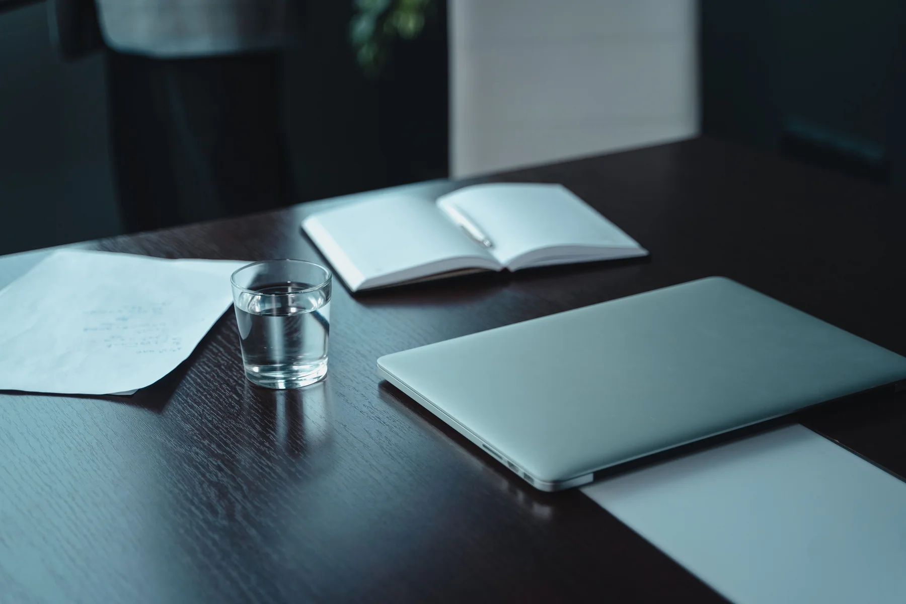 Ein geschlossener Laptop, ein Notizbuch und ein Glas Wasser auf einem dunklen Tisch.