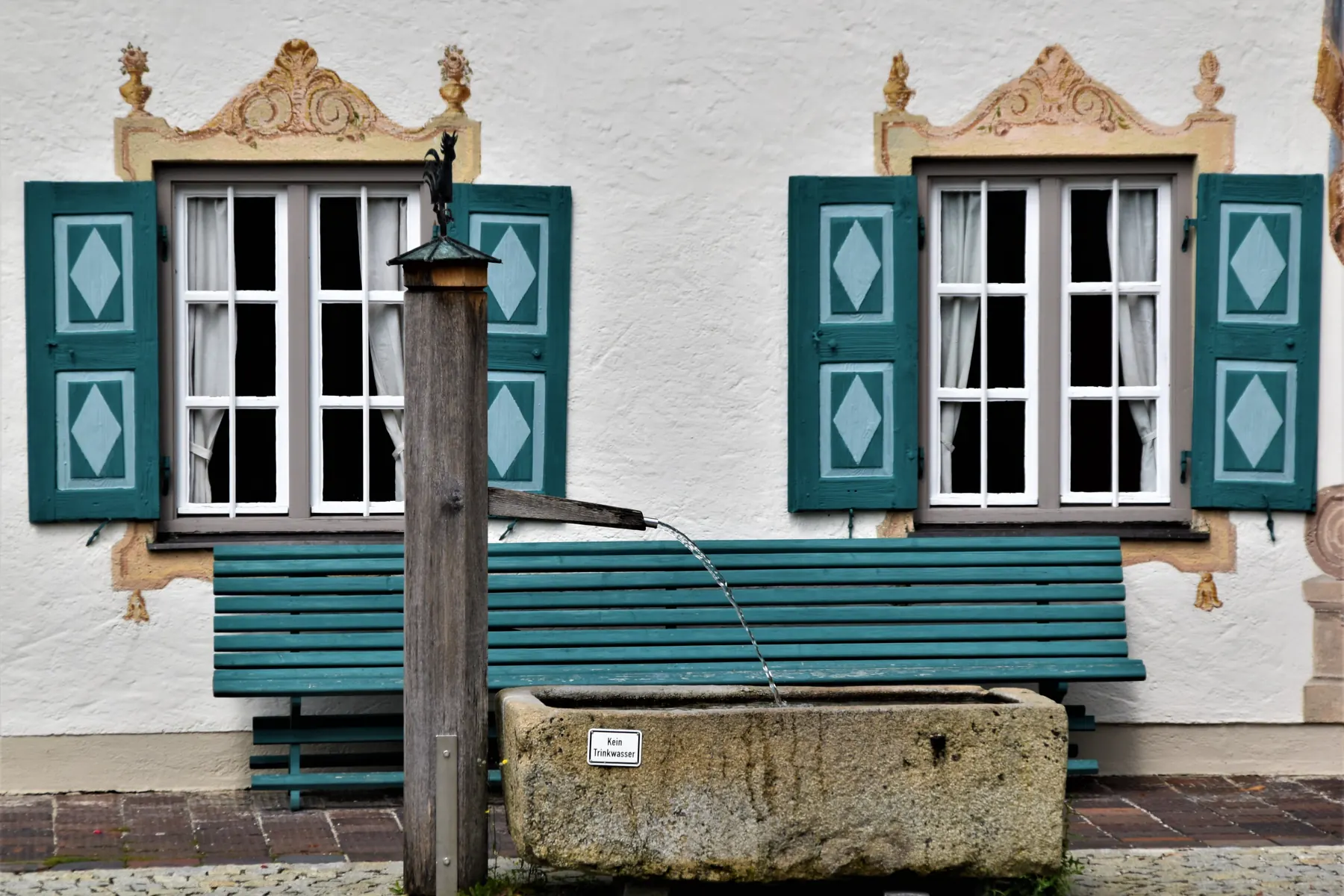 Ein weißes Haus mit grünen Fensterläden, davor ein Brunnen und eine Holzbank.